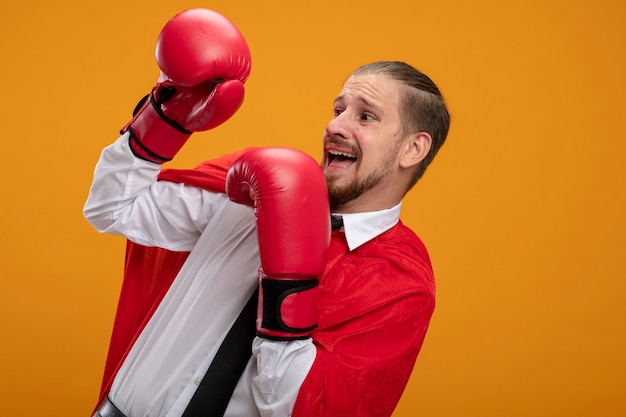 Scared young superhero guy wearing tie and boxing gloves looking at side covered face isolated on orange background