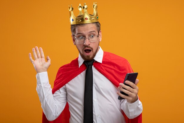 Scared young superhero guy wearing tie and crown with glasses holding and looking at phone and points with hand at side isolated on orange background