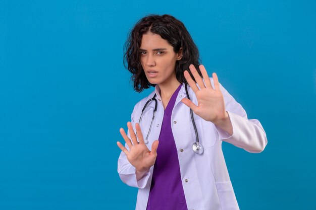 Испуганная молодая женщина-врач в белом халате со стетоскопом держит руки вверх и говорит, что не подходите ближе на изолированном синем