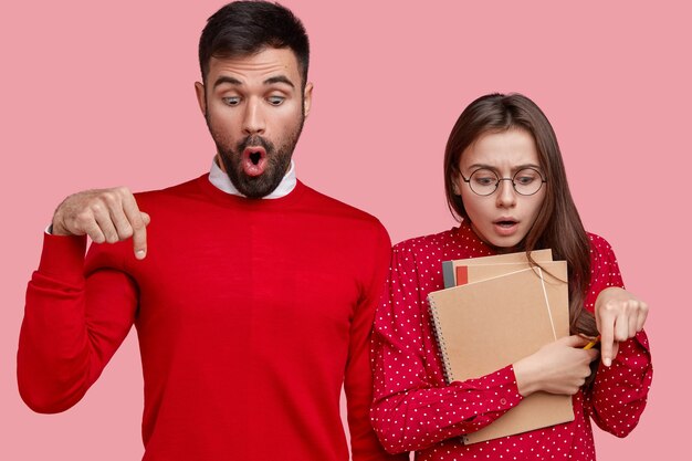 Scared young woman and man with terrified expression point down on floor, have puzzled look, dressed in red outfit