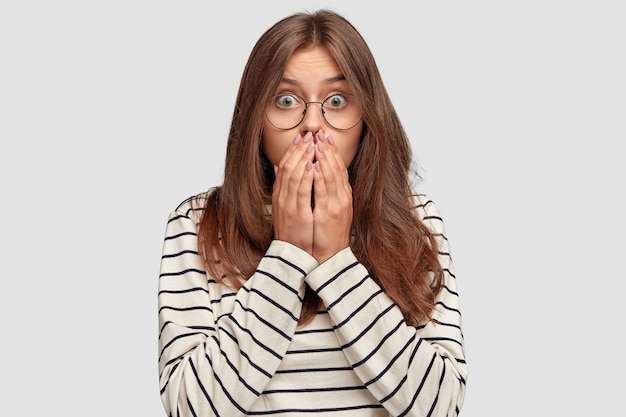 Scared young woman with glasses posing against the white wall