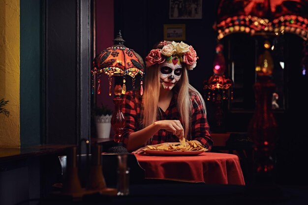 Scary blonde girl with undead makeup in flower wreath eating nachos at a mexican restaurant. Halloween and Muertos concept.