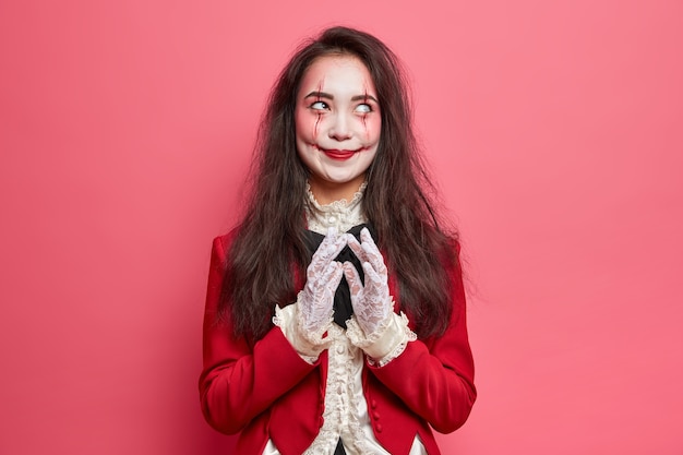 Scary brunette woman with zombie eyes and bloody scars dressed in halloween costume steepls fingers and has evil plan isolated over pink wall