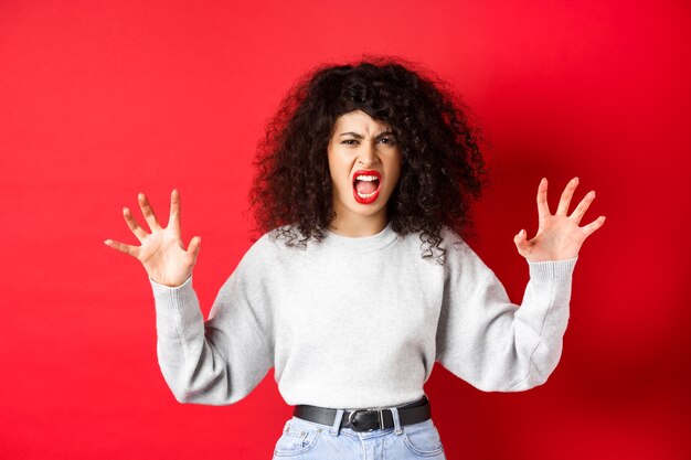 Scary woman trying to scare you, shouting and showing animal claws gesture, screaming at person, standing against red background.