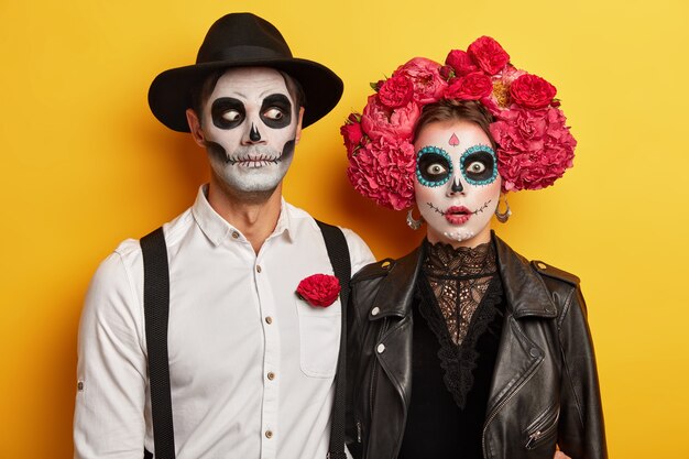 Scarying undead couple dressed in carnival costume, wears skull makeup, red flowers as symbol of this event.