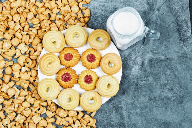 Scattered alphabet crackers, a plate of cookies and a jar of milk on marble table.