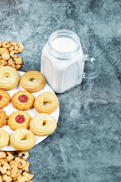 Scattered alphabet crackers, a plate of cookies and a jar of milk on marble table.