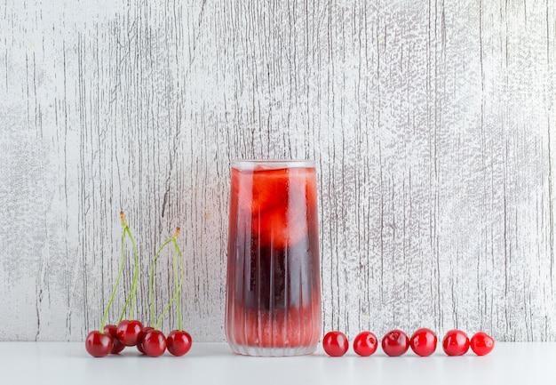 Scattered cherries with icy drink on white and grungy table, side view.