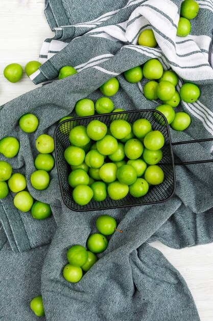 Scattered green plums in a black cullender on a kitchen towel and white wood. flat lay.