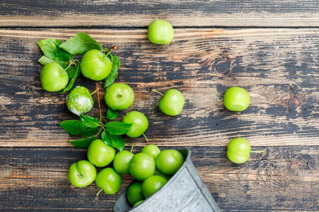 Scattered green plums from mini bucket with leaves, salt on wooden wall, top view.