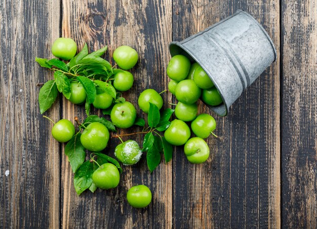 Scattered green plums with leaves, salt in a mini bucket on wooden wall, top view.