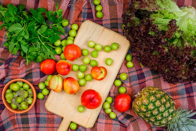 Scattered greengages and peaches with green leaves, a pineapple and lettuce in a cutting board and clay bowl on picnic cloth, flat lay.