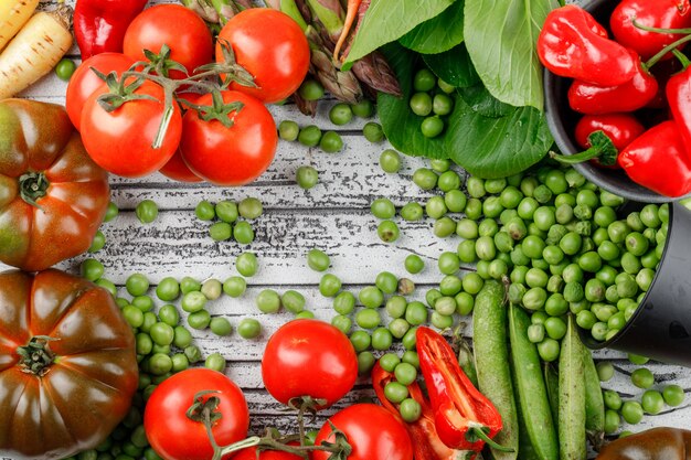 Scattered peas from a bucket with peppers, tomatoes, bok choy, green pods, asparagus, carrots flat lay on a wooden wall