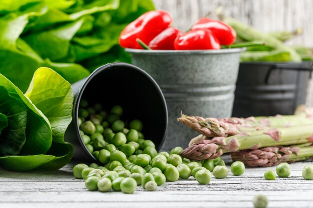 Scattered peas with lettuce, green pods, asparagus, peppers from a bucket on wooden wall, side view.