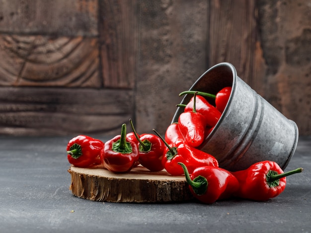 Scattered red peppers from a mini bucket with wooden piece side view on grey and stone tile wall