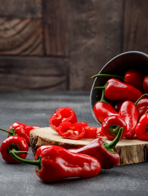 Scattered red peppers with wooden piece in a mini bucket on grey and stone tile wall, high angle view.