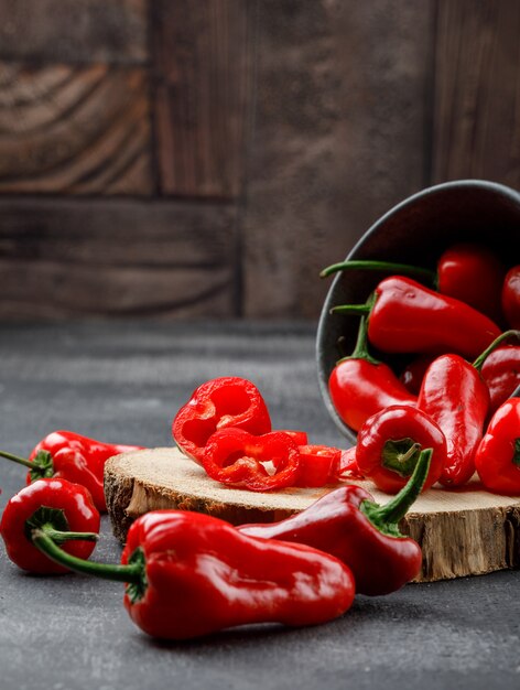 Scattered red peppers with wooden piece in a mini bucket on grey and stone tile wall, high angle view.