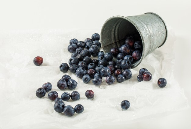 Scattered wet blueberries in a mini bucket with wrapping paper high angle view on a white surface