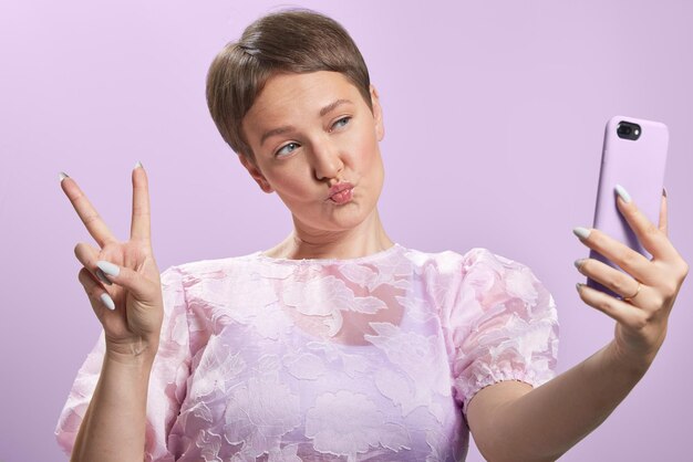 Scene young caucasian short hair woman make duck face or victory sign to make a selfie with her phone Puffy pink dress Isolated medium shot studio portrait on a purple background High quality photo