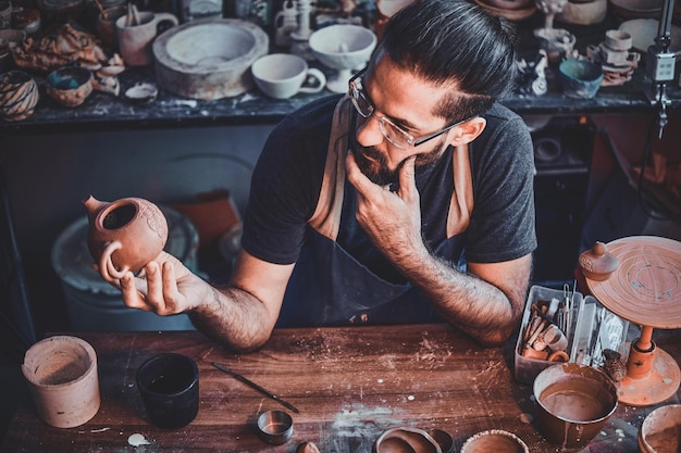 Sceptical hardworking man is looking at his own work, ceramic teapot.