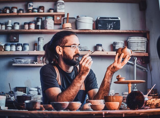 Sceptical hardworking man is looking at his own work, ceramic teapot.