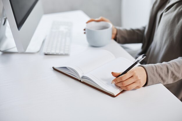 Schedule helps maintain my day. Cropped shot of woman working in front of computer, writing in notebook and drinking coffee. Businesswoman makes plan of her meeting during day