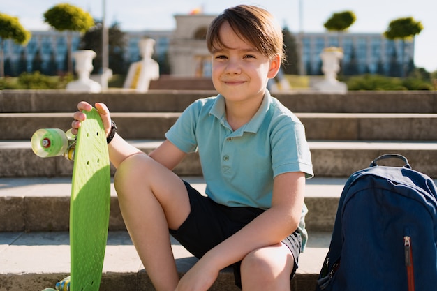 Foto gratuita school boy in polo blu seduto sulle scale con uno zaino blu e penny verde a bordo