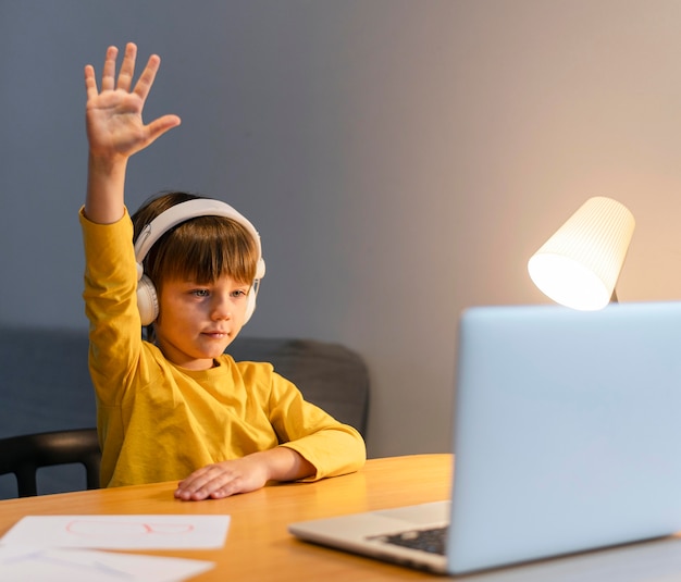 School boy in yellow shirt taking virtual classes and raising hand