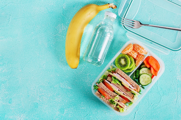 샌드위치, 야채, 물, 과일 테이블에 학교 점심 상자.