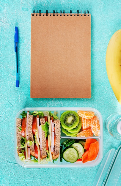Коробка школьного обеда с бутерброд, овощи, вода и фрукты на столе.