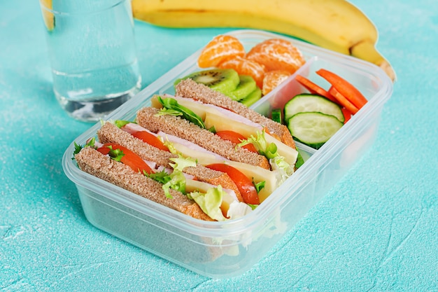 School lunch box with sandwich, vegetables, water, and fruits on table.