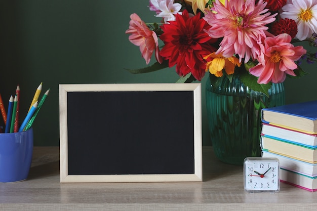 School mockup with an empty frame, a stack of textbooks, a bouquet of dahlias, pencils and an alarm clock on the table. back to school. september 1. teacher's day.