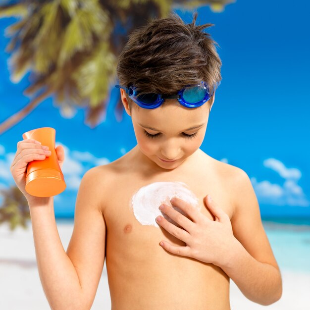 日焼けした体に日焼け止めクリームを塗る小学生の男の子。オレンジ色の日焼けローションボトルを持っている少年。