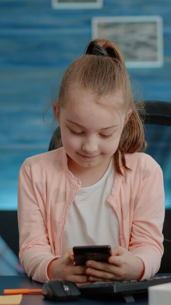 Schoolgirl looking at smartphone for homework and courses at home. Little child using mobile phone with touch screen for online remote class lessons and education at desk. Pupil learning