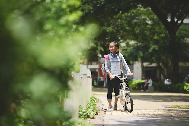 Школьница с рюкзаком гуляет на свежем воздухе со своим велосипедом после школы