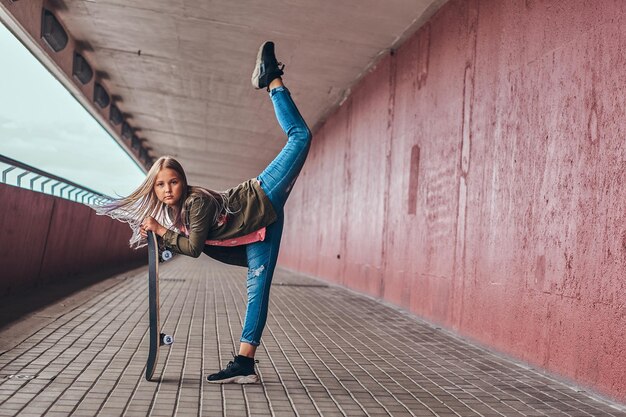 Schoolgirl with blonde hair dressed in trendy clothes dance with a skateboard at a bridge footway.