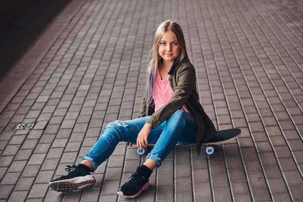 Schoolgirl with blonde hair dressed in trendy clothes sitting on a skateboard at a bridge footway.