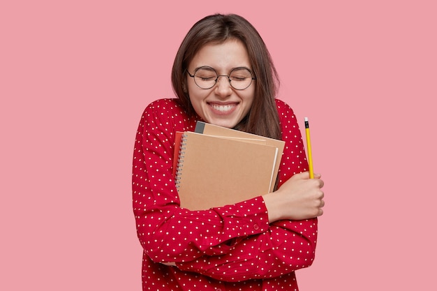 Schoolgirl with positive expression carries notebook closely to herself, smiles broadly, holds pencil, wears fashionable red shirt, isolated over pink background