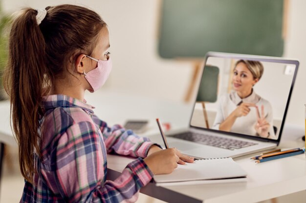 Schoolgirl writing while having online class with her teacher over laptop due to virus pandemic