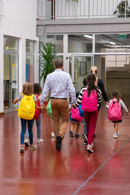 Школьники с яркими рюкзаками идут по школьному коридору, держась за руки учителей