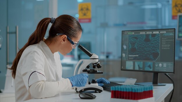 현미경을 사용하여 실험실에서 DNA 샘플을 분석하는 과학 엔지니어. 보호용 고글과 장갑을 끼고 실험실 도구와 컴퓨터에서 미세한 유리 렌즈로 작업하는 여성 생물학자.