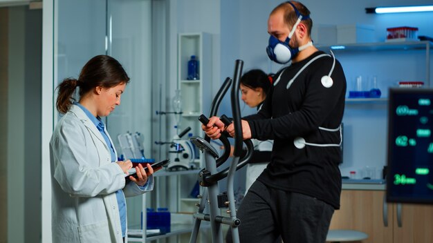 体にマスクと電極を取り付けたクロストレーナーで走っているスポーツマンが患者に健康について尋ねる科学スポーツ医師
