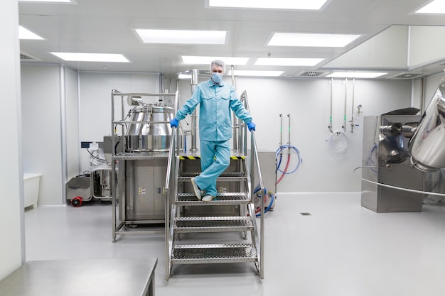 Ученый в синем лабораторном костюме стоит на металлической хромированной лестнице со скрещенными ногами