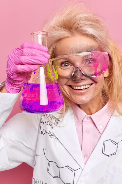  scientist exmaines pink and purple liquid in flask does scientific research wears protective glasses white coat works as laboratory assistant smiles pleasantly poses indoor