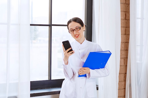 Scientist female holding folder and mobile phone near window .