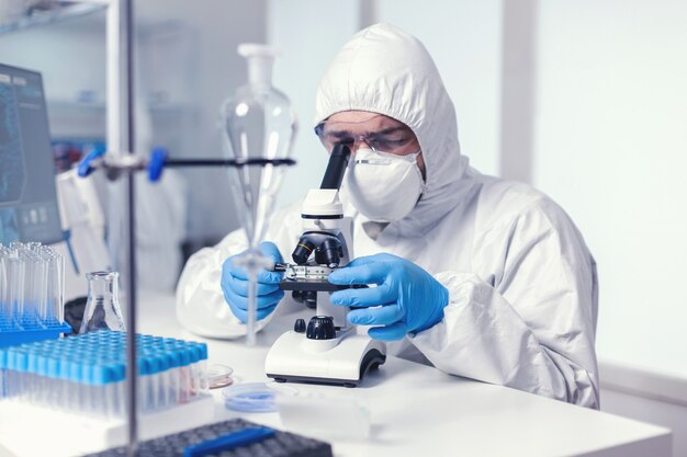 과학자는 그의 직장에 앉아 있는 양복을 입고 현미경을 들여다보고 있습니다. 코로나바이러스 발병 동안 작업복을 입은 Virolog가 의료 과학 분석을 수행합니다.