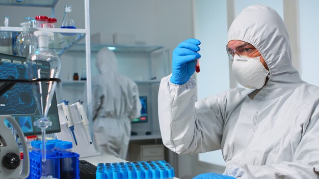 Scientist in sterile chemistry suit analysing blood sample from test tube. Viorolog researcher in professional lab working to discover medical treatment against covid19 virus.