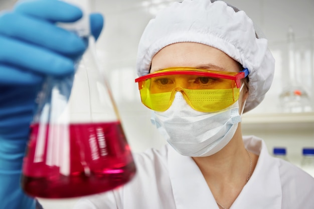 Scientist working in the lab, in protective mask and cap, examines a test tube with liquid