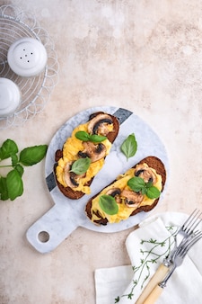 흰색 테이블 배경에 빵에 튀긴 버섯과 바질을 넣은 스크램블 에그. 집에서 만든 아침 식사 또는 브런치 식사. 스크램블 에그와 버섯 샌드위치. 복사 공간이 있는 상위 뷰