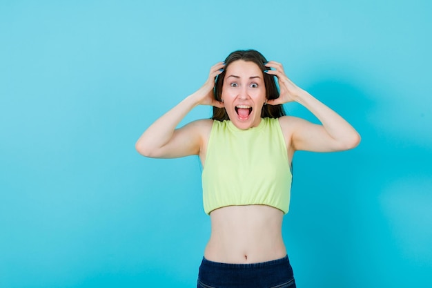 Free photo screaming girl is looking at camera by holding hands around head on blue background